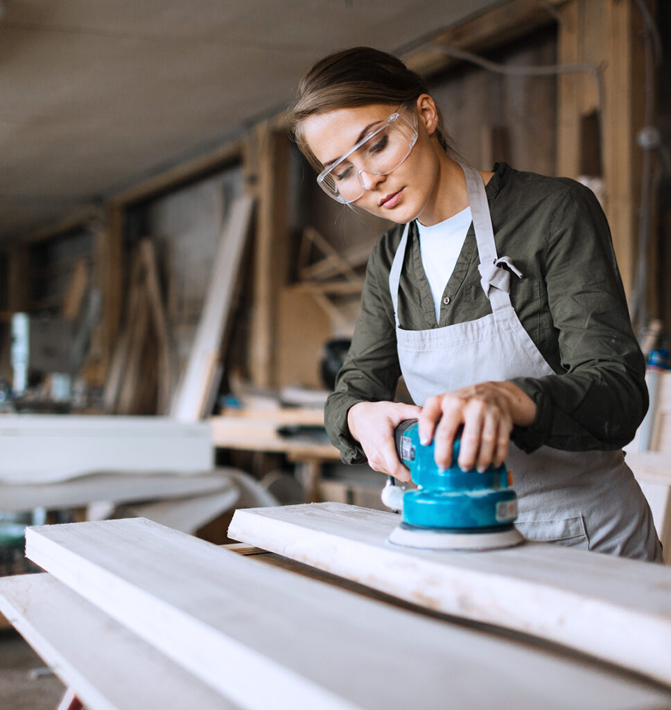 Eine junge Frau arbeitet in einer Werkstatt. Sie trägt eine Schutzbrille und schleift mit konzentriertem Blick die Oberfläche eines Holzbretts ab.