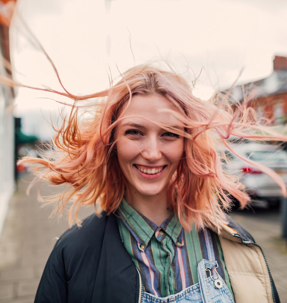 Eine junge Frau mit rosa Haaren steht draussen in einer Strasse. Ihre Haare wehen stark um ihr Gesicht, sie lächelt dabei direkt in die Kamera.