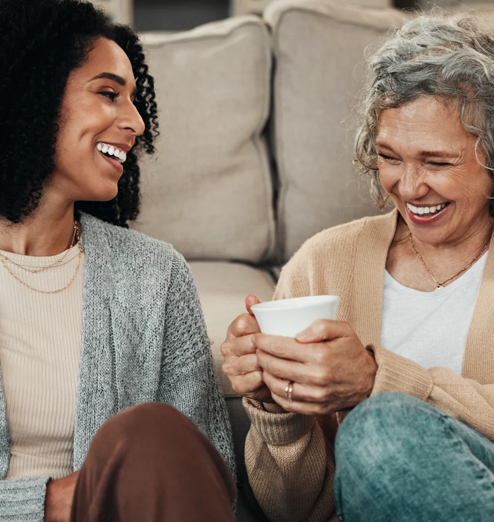 Eine jüngere Frau und eine Frau im mittleren Alter sitzen gemeinsam auf dem Boden vor einem Sofa und trinken Kaffee. Beide lachen herzlich.