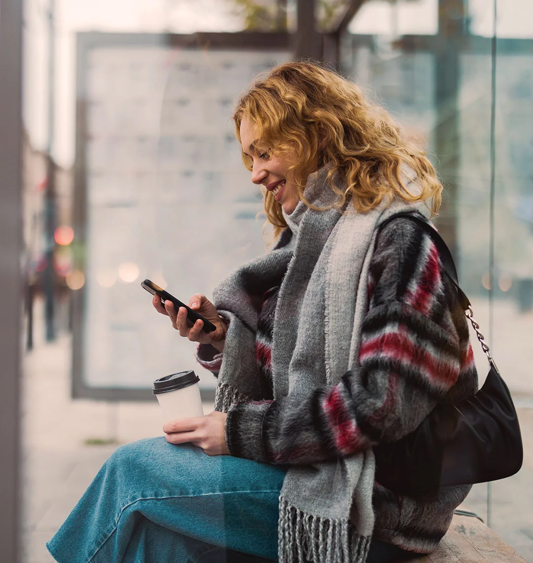 Eine junge Frau sitzt an einer Bushaltestelle und schaut lächelnd in ihr Handy.