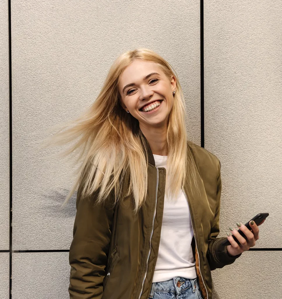 Eine blonde Frau lächelt in die Kamera und hält dabei ein Smartphone in der Hand.