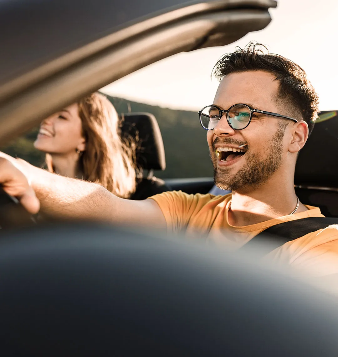 Ein junger Mann fährt mit seiner Beifahrerin in einem Cabrio und lacht dabei.