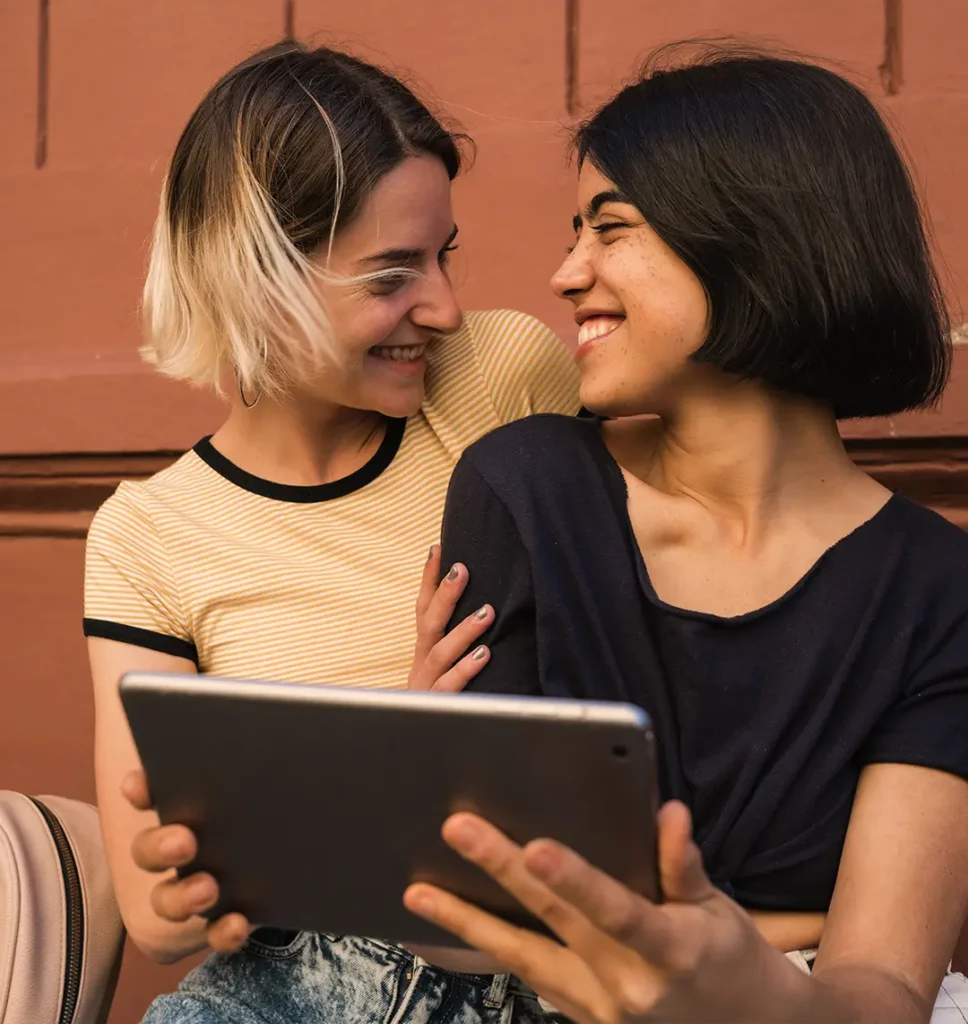 Zwei Frauen sitzen vor einem Tablet und schauen sich dabei gegenseitig an.