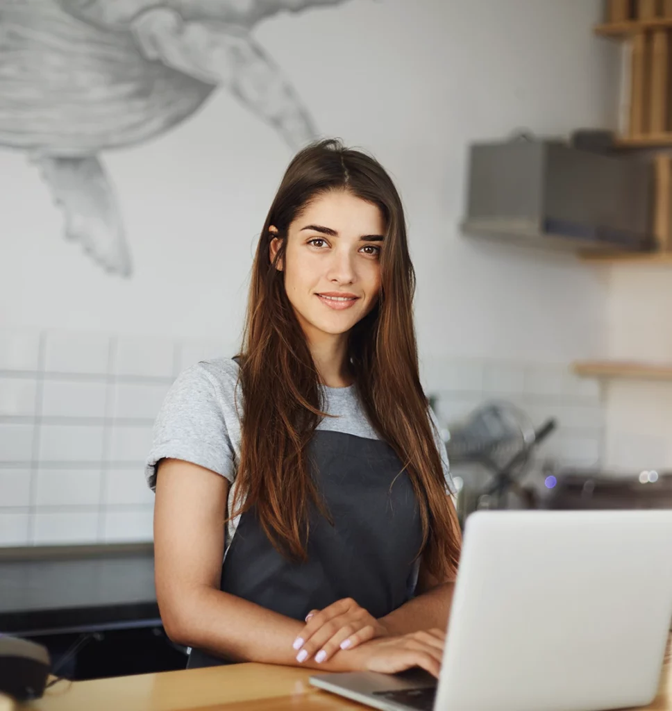 Eine junge Frau, die in einem Café arbeitet, steht hinter einem Tresen. Vor ihr steht ein Laptop. Sie trägt eine Schürze und lächelt in die Kamera.