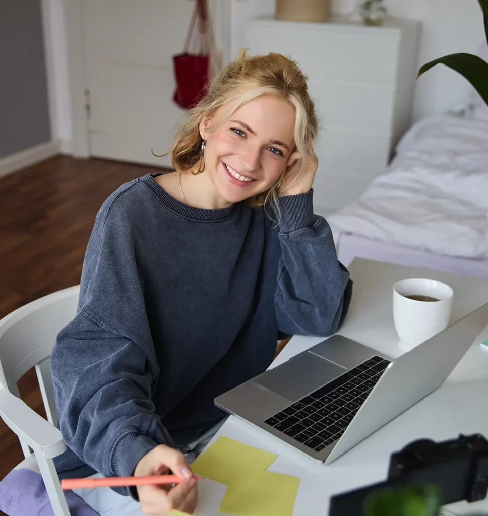 Eine Frau sitzt am Schreibtisch vor ihrem Laptop und lacht in die Kamera