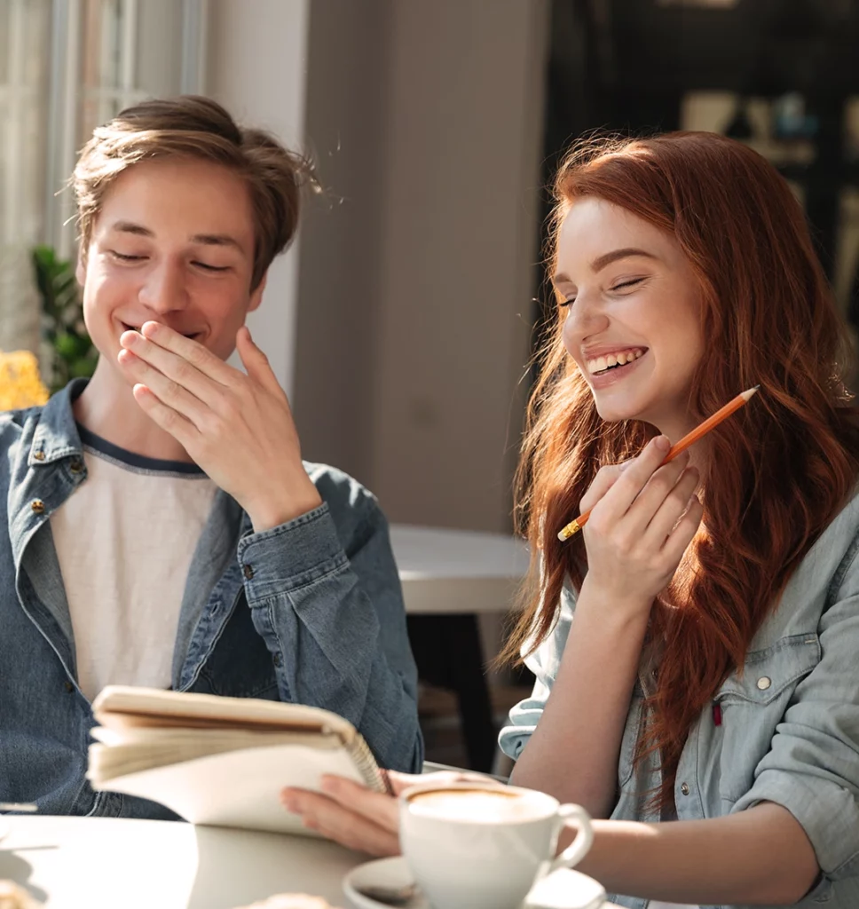 Ein Junge und ein Mädchen sitzen in einem Café und lachen. Sie hält einen Block und einen Stift in der Hand.