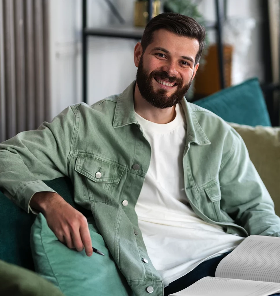 Ein Mann sitzt auf einem Sofa mit grünen Kissen in einem Wohnzimmer. Im Hintergrund steht ein schwarzes Regal mit einer Pflanze. Er schaut direkt in die Kamera und lächelt.Auf dem Schoß hat er ein Buch, in das man schreiben kann und in der Hand einen Stift.